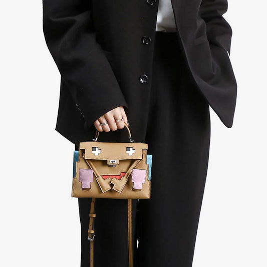 Lifestyle shot of a model carrying the Inspired Kelly Doll Picto handbag in brown Epsom leather, styled as a chic hand-carry mini tote against a black suit.