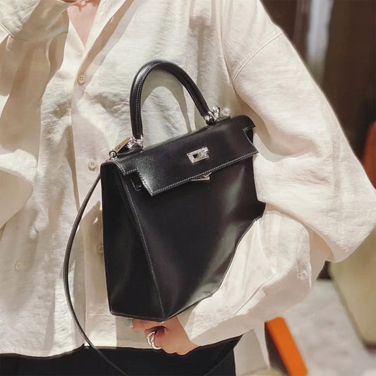 A person wearing a white linen shirt carrying a black Box Leather Kelly-style handbag with silver hardware and a long shoulder strap.