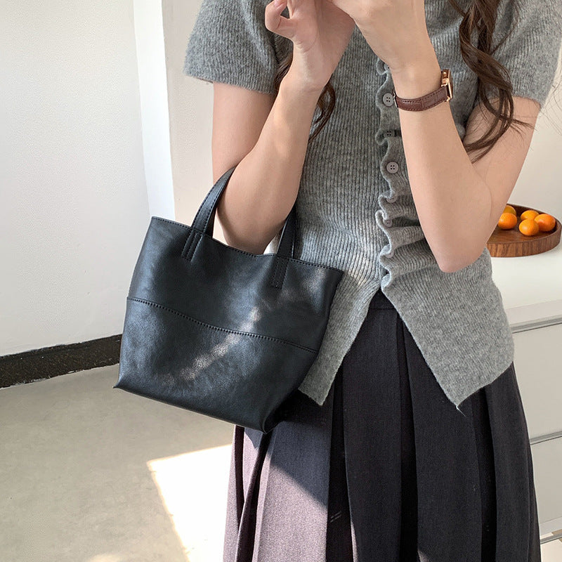 Minimalist black premium cowhide mini tote bag held by hand, styled with a grey knitted cardigan and black pleated skirt.