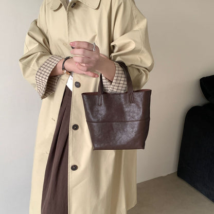 A mini tote bag crafted from premium dark brown cowhide, carried by a woman wearing a beige trench coat paired with a brown pleated skirt, presents a classic autumn look.