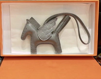 Gray lambskin Pegasus charm with crocodile-pattern wings in an orange gift box.
