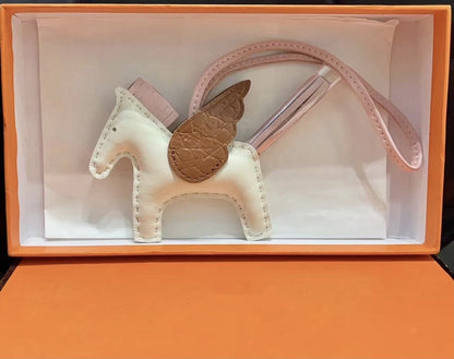 Tricolor Pegasus charm with off-white body, pink strap, and brown crocodile-pattern wings in an orange gift box.