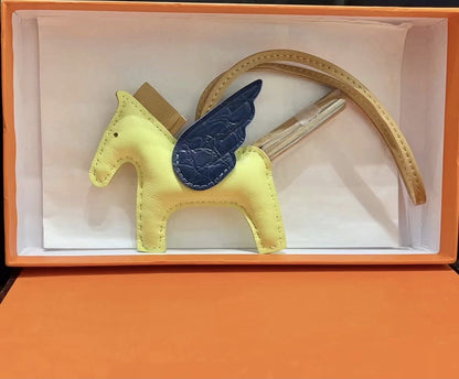 Tricolor Pegasus charm with light yellow body, navy blue crocodile-pattern wings, and tan strap in a gift box.