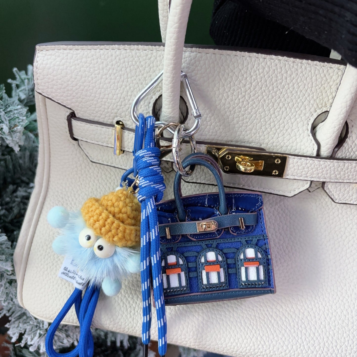 Blue Faubourg style mini Birk bag charm and earphone storage case made of vegan leather, styled on a cream handbag with a cute monster keychain.