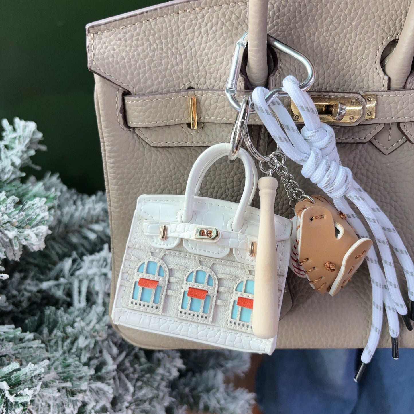 A white vegan leather Faubourg style mini Birk bag charm with light blue and orange window details, used as an earphone storage bag on a taupe handbag.