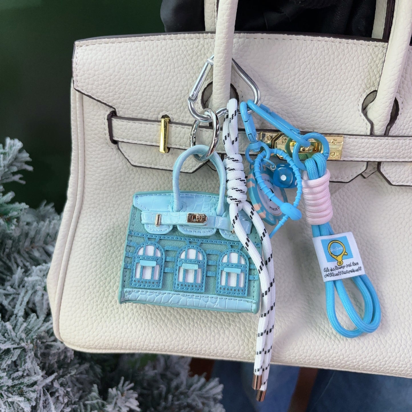 A light blue vegan leather Faubourg style mini Birk bag charm with white window details, styled as an earphone case on a white handbag with matching blue accessories.