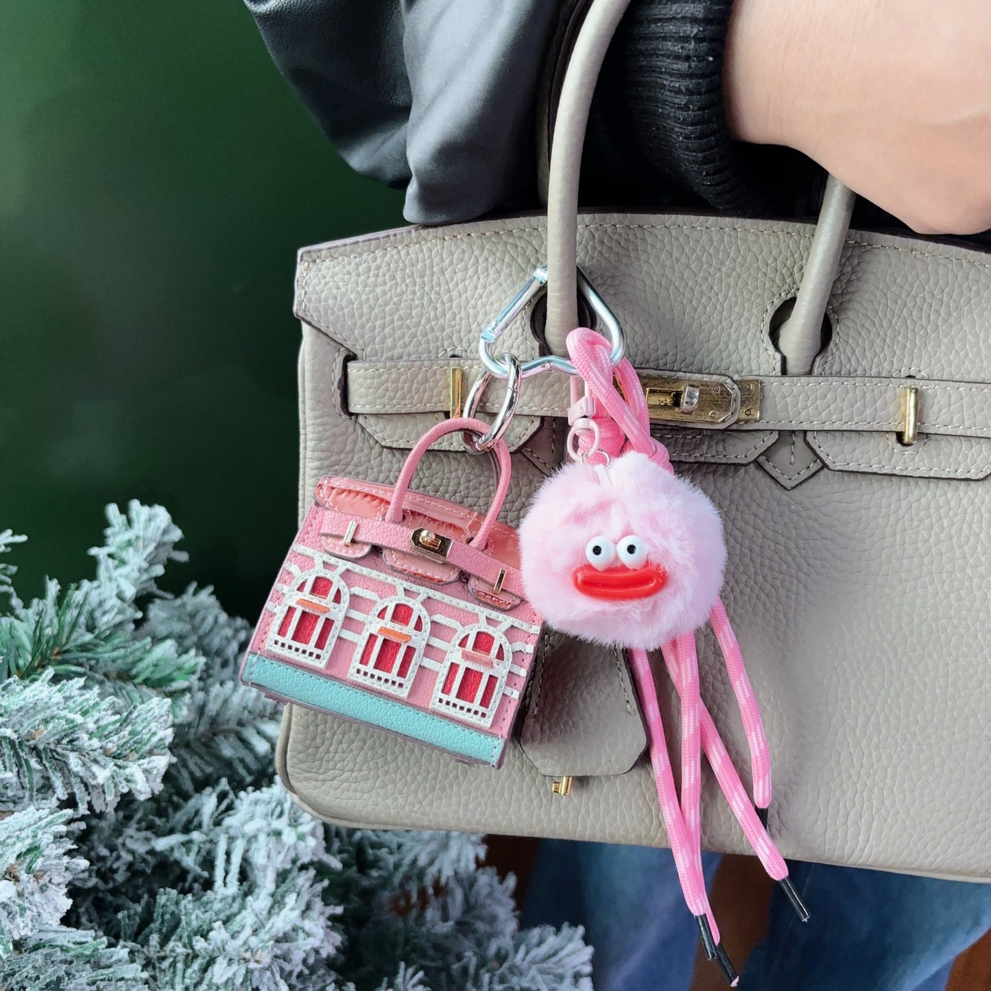 A pink and light blue vegan leather Faubourg style mini Birk bag charm with white window details, styled as an earphone case on a grey handbag with a fluffy pink monster charm.