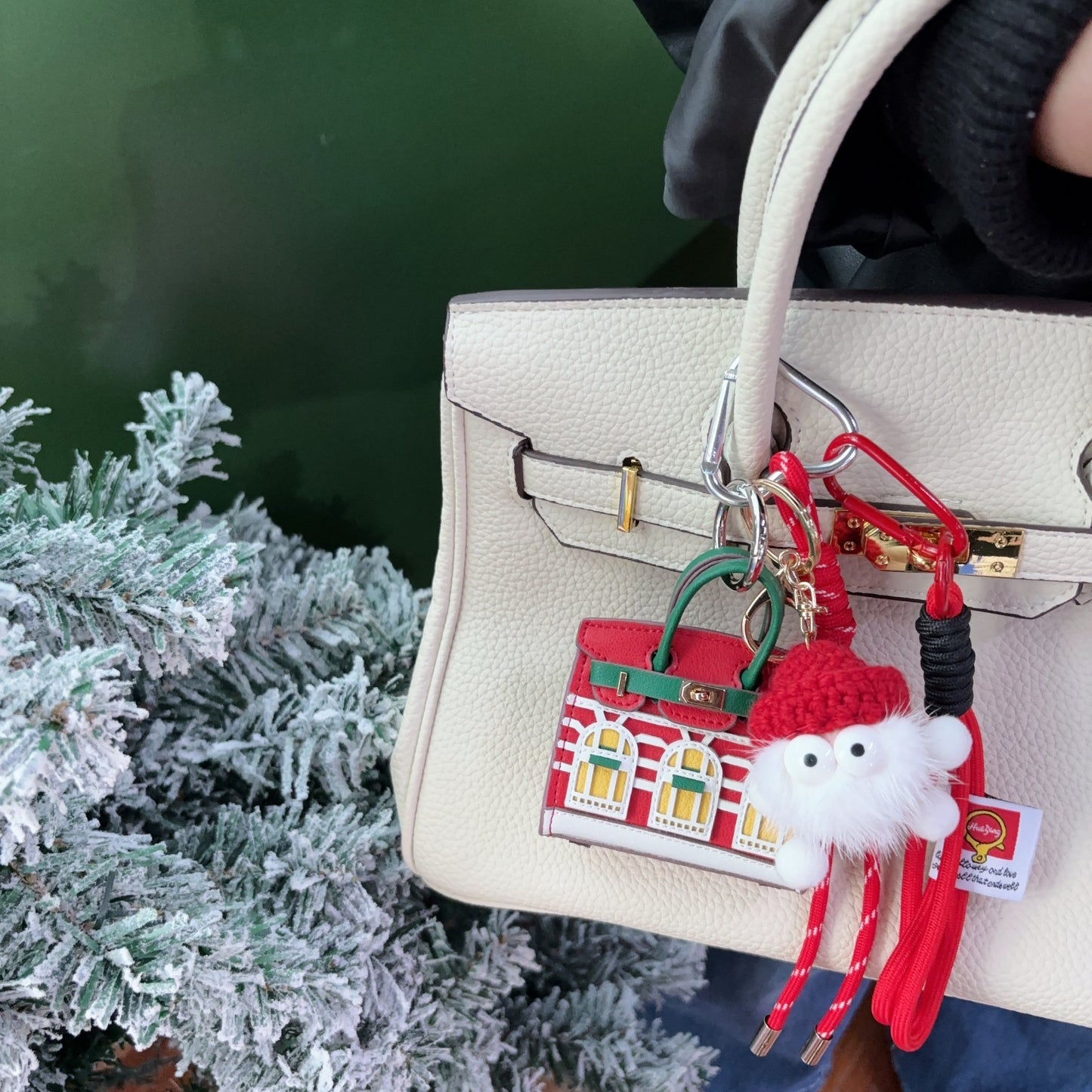 A festive red and green vegan leather Faubourg style mini Birk bag charm with white window details, styled as an earphone case on a cream handbag with a cute white monster keychain.
