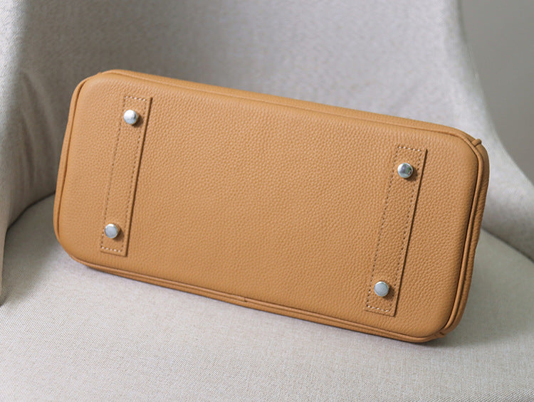 Bottom view of a tan JPG Birkin inspired handbag, showcasing four protective silver-tone metal feet and reinforced top grain cowhide leather base.