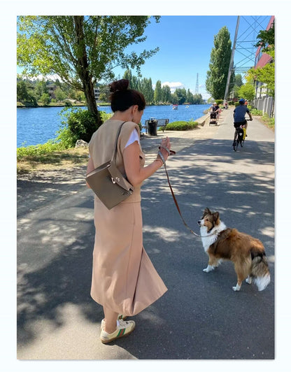 A woman wearing a taupe Kelli Danse top grain leather shoulder bag while walking her dog by a river, showing a relaxed casual lifestyle.