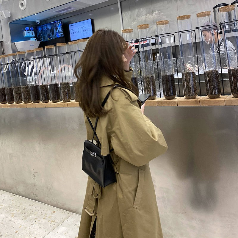 A woman styling a black top grain leather Kelli Danse bag as a backpack over a classic khaki trench coat in a modern cafe setting.