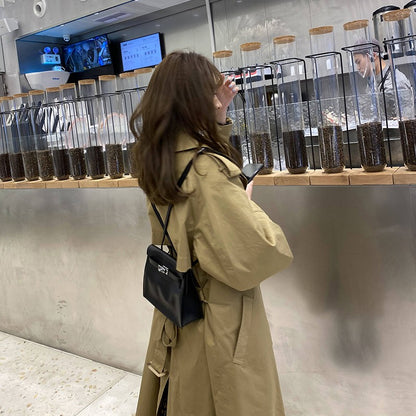 A woman styling a black top grain leather Kelli Danse bag as a backpack over a classic khaki trench coat in a modern cafe setting.
