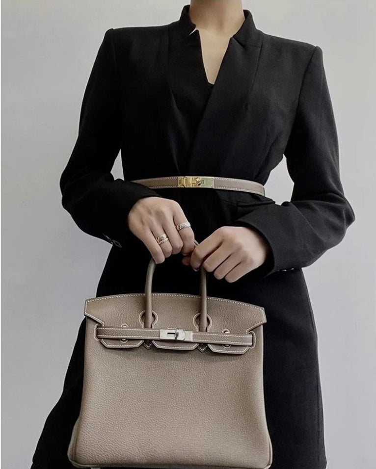 A woman wearing a black blazer styled with a taupe Epsom Kelli belt and a matching taupe leather Birkin handbag.