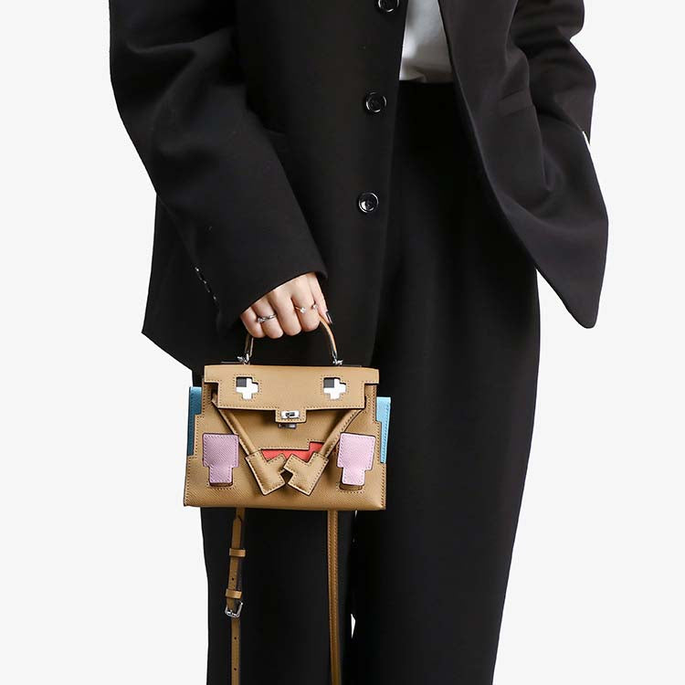 Lifestyle shot of a model carrying the Inspired Kelly Doll Picto handbag in brown Epsom leather, styled as a chic hand-carry mini tote against a black suit.