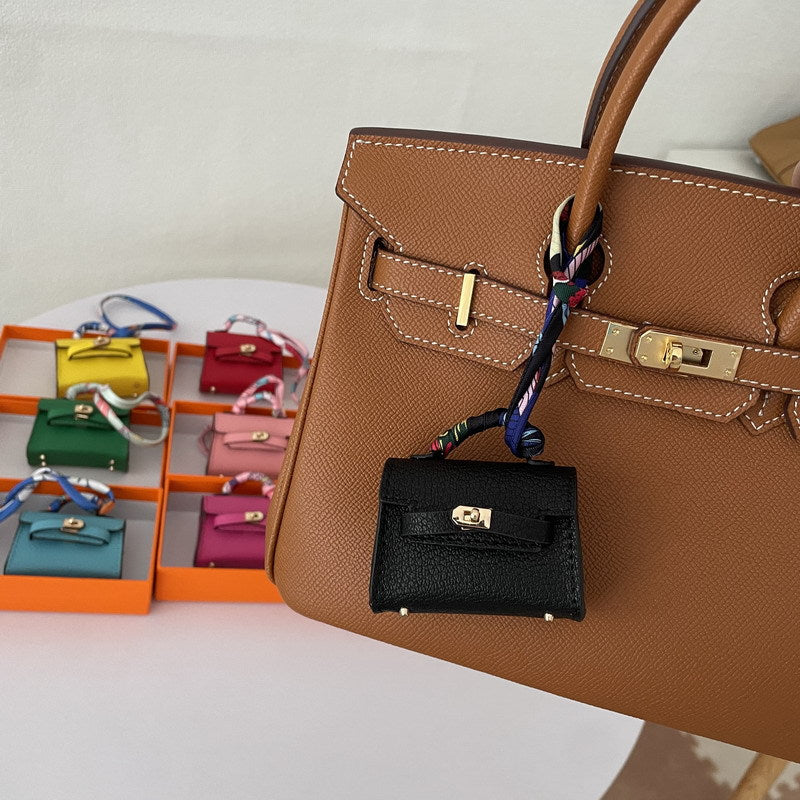 A black Mini Kelly bag charm, adorned with a cascading silk scarf detail, suspended from a brown tote bag.
