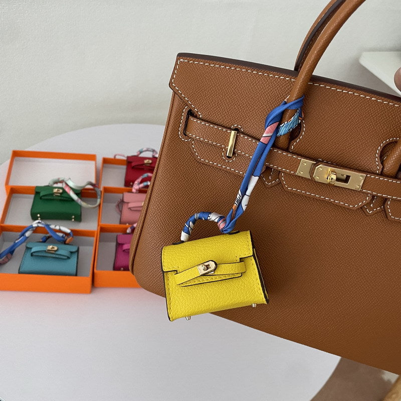 A bright yellow Mini Kelly bag charm, adorned with a blue silk scarf, hangs from a brown bag.