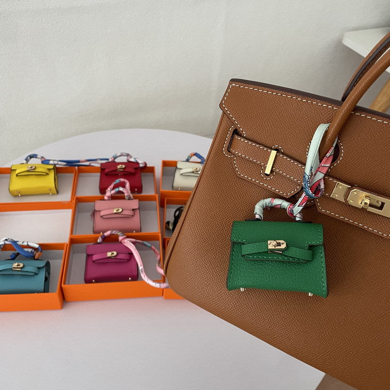 A green mini Kelli bag charm, adorned with a light-colored silk scarf, hangs from a brown tote bag.