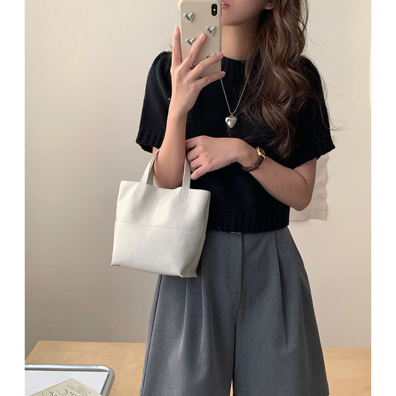 Minimalist white premium cowhide mini tote bag held by hand, styled with a black short-sleeve knit top and grey high-waisted trousers.