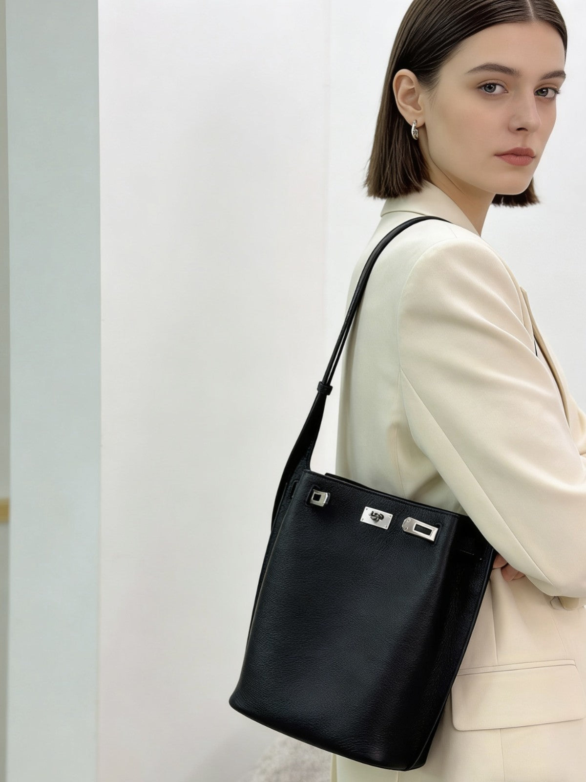 Model wearing a black pebbled top-grain leather Kelly bucket bag as an underarm shoulder bag with a cream blazer.