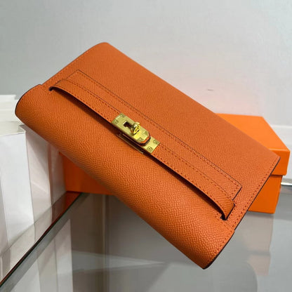 Angled view of an Orange inspired Kelly to go wallet in textured cowhide leather, featuring the iconic golden Kelly closure and signature orange packaging in the background.
