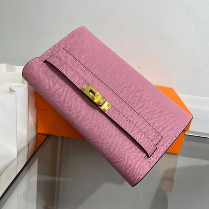 Angled view of a pink inspired Kelly to go wallet in textured cowhide leather, featuring the iconic golden Kelly closure and signature orange packaging in the background. 